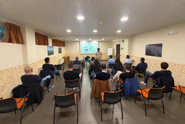 Foto reale ritoccata: partecipanti visti dal fondo dell’aula seguono una spiegazione del docente davanti allo schermo.